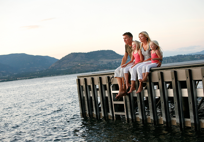 family of 4 wearing white pants, girls wearing coral shirts, sitting on a dock by the lake and mountains