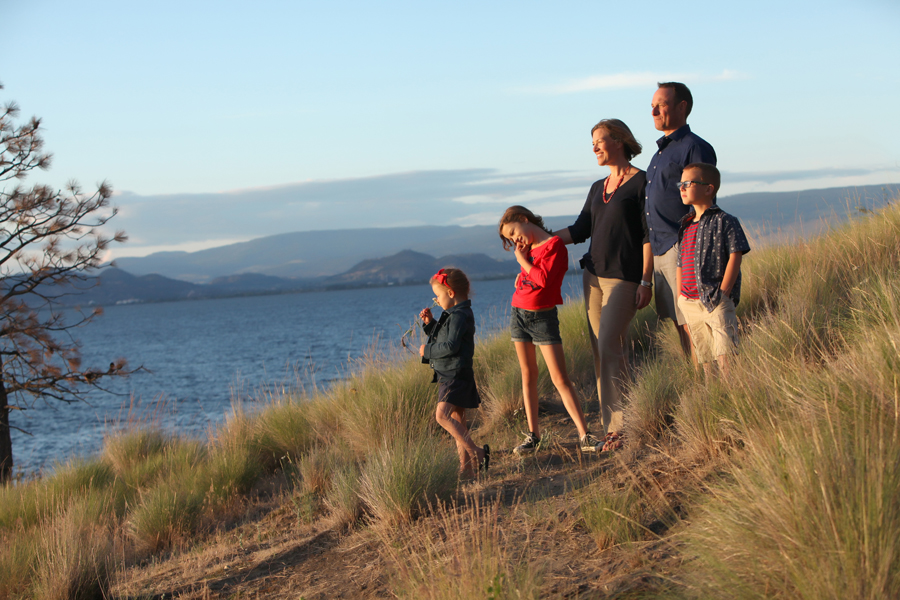 Photo of family of 5 standing on a hill of dried grass by lake and mountains