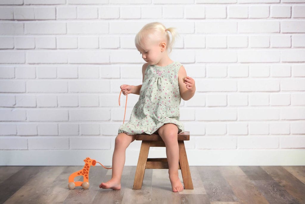 Young girl with blond pig-tails wearing a green dress playing with an orange toy sitting on a stool infant of white brick