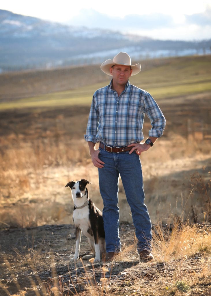Business Portrait of a man wearing blue plaid and a white cowboy hat standing in a field of dried grass and mountains with dog