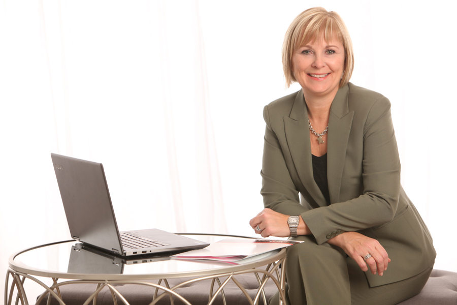 Business Portrait of a blond woman wearing an olive suit sitting by a table with laptop in front of white curtains