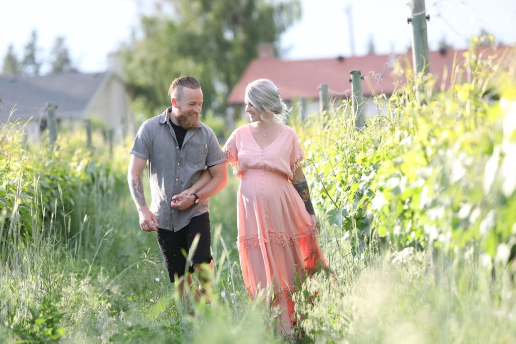 Maternity Portrait of a couple wearing a pink dress and grey shirt walking together down an Okanagan vineyard row with houses