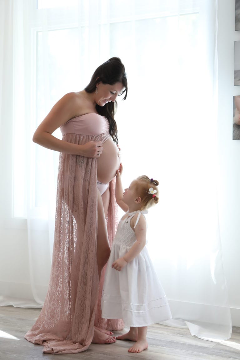 Maternity Portrait fo a pregnant mother wearing pink fabric and standing with toddler wearing a white dress in front of window with white curtains