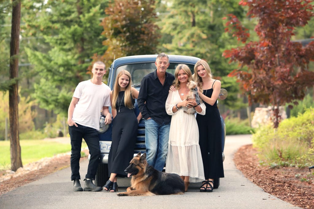 family of 5 wearing black, blue and white standing with their dog in front of a vintage truck on a driveway with trees