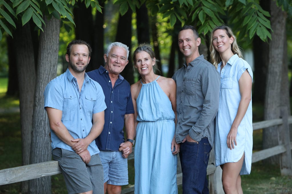 family of 5 wearing grey and blue stand and sit by a rustic fence by forest