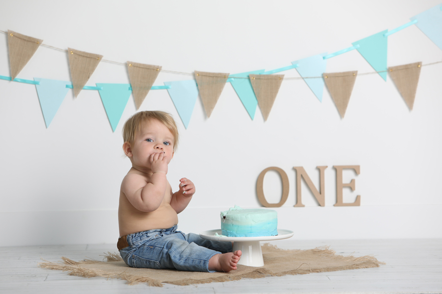 one year old boy wearing jeans and eating cake on a burlap mat in front of aqua coloured pennants