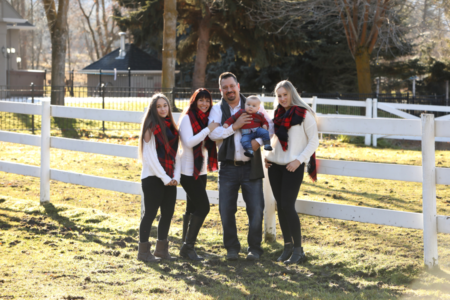 family of 5 wearing white and red plaid hold baby boy in front of a white horse fence