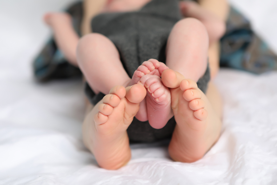 Sibling holds newborn on lap, photograph is taken as a close-up of their feet with dark green blankets