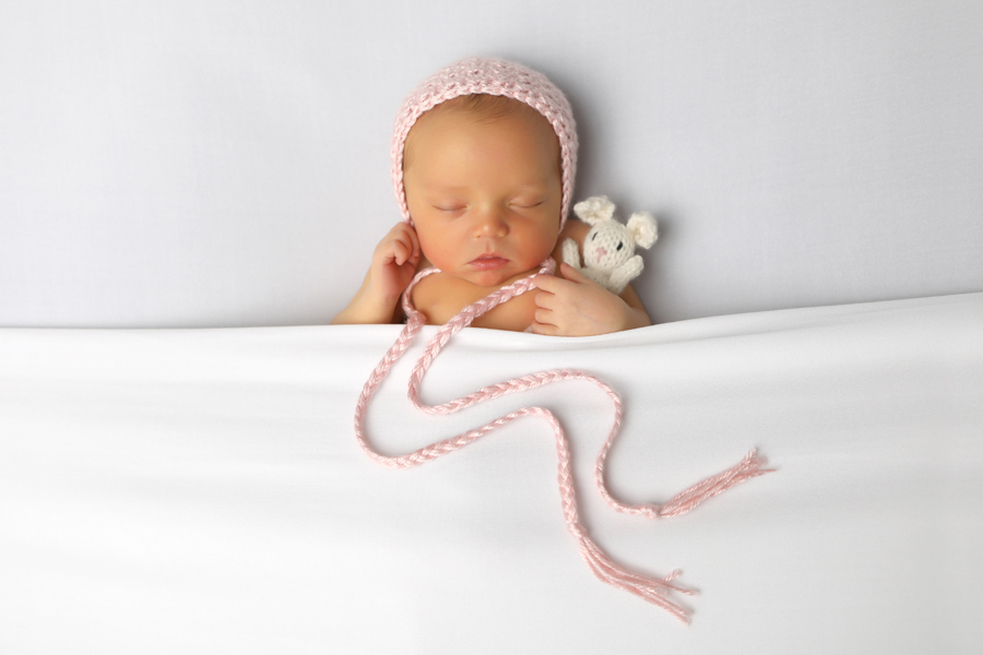 Newborn wearing a pink bonnet sleeping while holding a tiny white bunny in white blankets