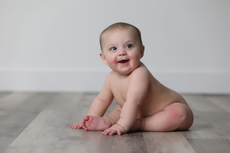 Smiling, naked baby seated on grey hardwood floors.