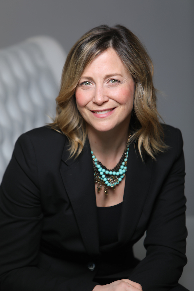 business headshot portrait of a woman in a black suit wearing a turquoise necklace against a grey background